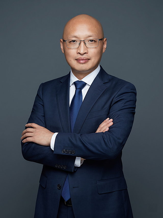 参考图中保留人物脸型、发型、年龄等特征生成职场形象照,标准焦段50mm,f/4光圈(f/2.8-f/4区间),平视视角(镜头与人物视线齐平);人物占画面70%,人物居中(居中构图),位于画面中心,为视觉聚焦点,入境范围头部及以下至臀部下方10cm(半身像);左右留少量背景空间;头部与画面顶部保留5%空间;头部发型完整,头肩比例协调,人物结构准确;发质微微柔顺，微微蓬松。身穿精纺羊毛混纺面料的深墨蓝色西装外套(含5%桑蚕丝提升柔光质感),平驳领(商务感正式感),左胸袋无口袋巾(简化高级感),西装下摆自然垂度;内搭长绒棉白色衬衫(温莎领),袖口贴合手腕1cm(无堆叠);搭配精致且高级的深蓝色斜向微微渐隐光泽感极细条纹商务感领带(丝提花材质,低饱和柔光渐隐);下装精纺羊毛混纺面料的深墨蓝色高腰直筒西裤(面料同西装);搭配黑色哑光头层牛皮皮带(宽2.5cm),带扣为长方形拉丝黑色金属(1.8cm×3cm) ;着装正式干练;专业稳重;身形匀称,姿态端正,身体挺拔。呈正面抱胸的自然站立姿态:左臂搭于右臂上方,肩颈自然松弛下沉,姿态自信从容;佩戴方形眼镜;简约冷调深灰色纯色背景(呈现均匀柔和质感),背景光线均匀柔和,强化色彩对比的高级感。极简干净;画质超高分辨率、细节清晰,现代精致感风格;肤质精细化处理;眼神直视镜头,目光坚定且澄澈,表情自然。嘴角上扬;肤质自然细腻,肤色健康均匀,皮肤状态清爽干净,保留真实皮肤细节｡正面柔光补光,4800K中性光,面部光比1:1.5｡中近景写实摄影,对焦清晰,面部雾面柔光,轻透裸肌感,高清保留皮肤细节｡分辨率4K,去除人物之外的所有元素。严格按照图片内容生成。画面比例3:4