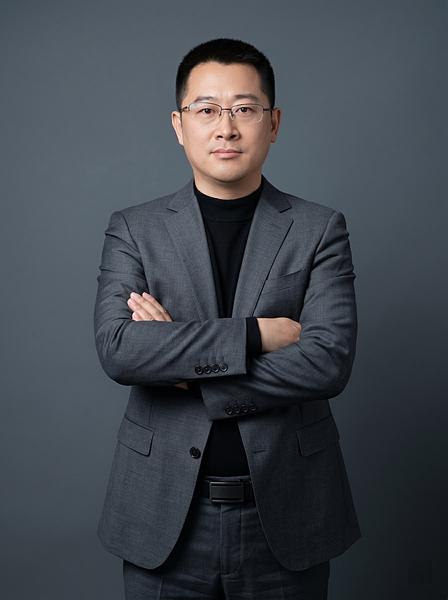 参考图中保留人物脸型、发型、年龄等特征生成职场形象照，标准焦段50mm,F/4光圈（处于F/2.8-F/4区间），平视视角（镜头与人物视线齐平）；人物占画面70%，人物居中（居中构图），位于画面中心，为视觉聚焦点，入境范围头部及以下至臀部下方10cm（半身像）构图。身穿修身精纺羊毛混纺面料的深灰色西装外套（含5%桑蚕丝提升柔光质感），平驳领（商务感正式感），左胸袋无口袋巾（简化高级感），西装下摆自然垂度；内搭黑色中领紧身针织上衣，下装精纺羊毛混纺面料的深灰色高腰直筒西裤（面料同西装），搭配黑色哑光头层牛皮皮带（宽1.5cm），带扣为长方形拉丝黑色金属（1.8cm×2.5cm）；着装正式干练，专业稳重；身形匀称，姿态端正，身体挺拔。呈正面抱胸的自然站立姿态，左臂搭于右臂上方，肩颈自然松弛下沉，姿态自信从容；佩戴细框眼镜。发梢微微修整，干练大气，主体锐利。简约冷调深灰色纯色背景（呈现均匀柔和质感），背景光线均匀柔和，强化色彩对比的高级感，极简干净；画质超高分辨率、细节清晰，现代精致感风格；肤质精细化处理；眼神直视镜头，目光坚定且澄澈，表情自然；肤质自然细腻，肤色健康均匀，哑光肤质清爽干净，保留真实皮肤细节；杰作，最佳画质，超高细节，写实摄影，中近景人像，皮肤质感自然清晰，柔光，正面补光，画面干净清新，高清皮肤细节；面部光比1:1.5，4800K中性光，微微修饰脸型，肤质哑光细腻，面部雾面柔光，轻透裸肌感，真实毛孔纹理；分辨率4K，严格按照图片内容生成，画面比例3:4