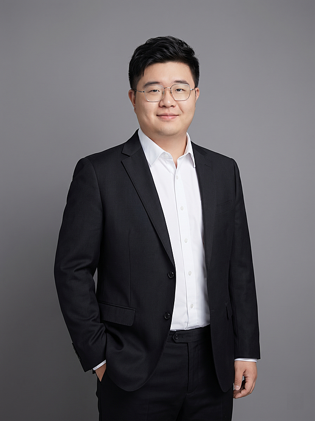 参考图中保留人物脸型、发型、年龄等特征生成教育行业职场形象照,标准焦段50mm,f/4光圈(f/2.8-f/4区间),平视视角(镜头与人物视线齐平);人物占画面70%,人物居中(居中构图),位于画面中心,为视觉聚焦点,入境范围头部及以下至臀部下方10cm(半身像);左右留少量背景空间;头部与画面顶部保留5%空间;头部发型完整,头肩比例协调,人物结构准确;发质微微柔顺，微微蓬松。身穿深黑色精纺羊毛混纺西装外套,(平驳领设计,面料呈细腻微微哑光质感);内搭白色高支棉衬衫;下装深黑色精纺羊毛混纺西裤;着装正式干练;专业稳重,博学温润;身形匀称,姿态端正,身体挺拔。呈站姿，左手插兜，肢体放松且自信;佩戴眼镜;简约微微中度灰色纯色背景(呈现均匀柔和质感),背景光线均匀柔和,强化色彩对比的高级感。极简干净;画质超高分辨率、细节清晰,现代精致感风格;肤质精细化处理;眼神直视镜头,目光坚定且澄澈,表情自然。嘴角上扬;肤质自然细腻,肤色健康均匀,皮肤状态清爽干净,保留真实皮肤细节｡正面柔光补光,4800K中性光,面部光比1:1.5｡中近景写实摄影,对焦清晰,面部雾面柔光,轻透裸肌感,高清保留皮肤细节｡分辨率4K,去除人物之外的所有元素。严格按照图片内容生成。画面比例3:4