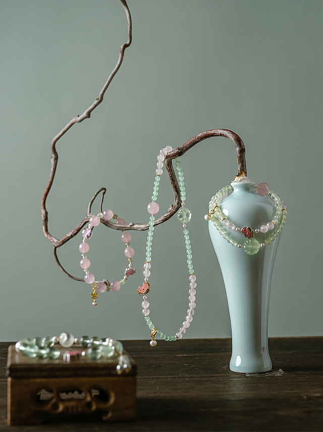 首饰（玉髓手链项链一套），严格参照上传第一张产品图，完整保留所有可见组件、部件、轮廓形状、结构、比例、材质质感、颜色，与上传产品图完全一致，不改变产品本身的颜色和细节。仅更换背景色，将原图棕色背景替换为与第二张参考图相似度50%的灰绿色调，保留产品原有展示结构和构图，保留枝干、青瓷瓶和木盒的位置和造型。整体是简约静物展示，背景为干净的莫兰迪灰绿色，柔和漫射顶光，产品受光面泛起灰绿色，暗部映射浅灰光晕，与环境物理融合。消除画面中多余的文字。4K分辨率，严格锁定宽高比为 3:4。