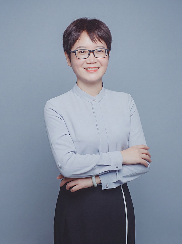 参考图中保留人物脸型、发型、年龄等特征生成金融法律行业职业形象照,标准焦段50mm,f/4光圈(f/2.8-f/4区间),平视视角(镜头与人物视线齐平);人物占画面70%,人物居中(居中构图),位于画面中心,为视觉聚焦点,入境范围头部及以下至臀部下方10cm(半身像);左右留少量背景空间;头部与画面顶部保留5%空间;头部发型完整,头肩比例协调,人物结构准确;发质微微柔顺，微微蓬松。身穿浅蓝高支精梳棉立领长袖衬衫;下装黑色西装半裙;着装正式干练;严谨理性、值得信赖;身形匀称,姿态端正,身体挺拔。呈自然站姿，双手抱胸，肢体放松且自信;佩戴黑框眼镜;佩戴精致玉镯配饰;简约冷灰蓝纯色背景(呈现均匀柔和质感),背景光线均匀柔和,强化色彩对比的高级感,极简干净;画质超高分辨率、细节清晰,现代精致感风格;肤质精细化处理;眼神直视镜头,目光坚定且澄澈,表情自然。嘴角上扬;清透自然裸妆(轻薄底妆+淡唇妆)妆感轻薄轻透裸肌感,通透感肤质,肤质自然细腻,肤色健康均匀,雾面柔光裸肌感,保留真实皮肤细节｡正面柔光补光,4800K中性光,面部光比1:1.5,色调明亮｡中近景写实摄影,对焦清晰,高清保留皮肤细节｡分辨率4K,去除人物之外的所有元素。严格按照图片内容生成。画面比例3:4