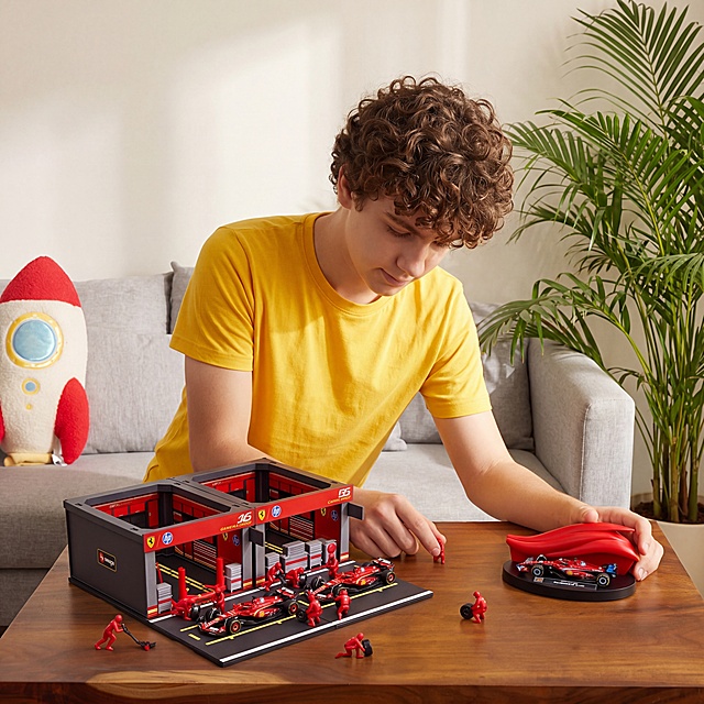 法拉利F1拼装玩具模型套装，严格参照上传产品图https://gdesign-dam-hz.dancf.com/36641627444336704/c25887fcd26f46ad9715b10221878657.webp，完整保留所有可见组件、部件、轮廓形状、结构、比例、材质质感、颜色与上传图完全一致。
室内家居桌面场景，暖棕自然色调。
白人青少年男孩俯身桌面摆弄玩具模型，保留参考图人物性别、国籍、肤色、年龄段、姿态、与产品互动方式，侧前方斜入柔和自然光，还原参考图光影方向，产品受光面泛起暖棕木色、暗部映射浅灰光晕，与环境物理融合。
产品居中偏前景，特写构图，微侧25°视角微调偏转。
全量换款清单：①人物：参考图浅蓝牛仔衬衫搭配酒红色长袖内搭 → 生成图亮黄色短袖T恤，保留卷发特征（性别、国籍、肤色、姿态保留，换服装+发型）；②背景置物架：参考图开放式原木置物架搭配瓶罐装饰 → 生成图浅灰色沙发搭配红白色火箭抱枕，右侧放绿色散尾葵盆栽；③桌面：参考图浅橡木色 → 生成图深胡桃木色；④保留孩子与玩具互动的动态感。
相似度50%，仅复刻参考骨架结构和氛围，不1:1复制。消除产品本体外多余品牌文字。
4K 分辨率，严格锁定宽高比为 1:1