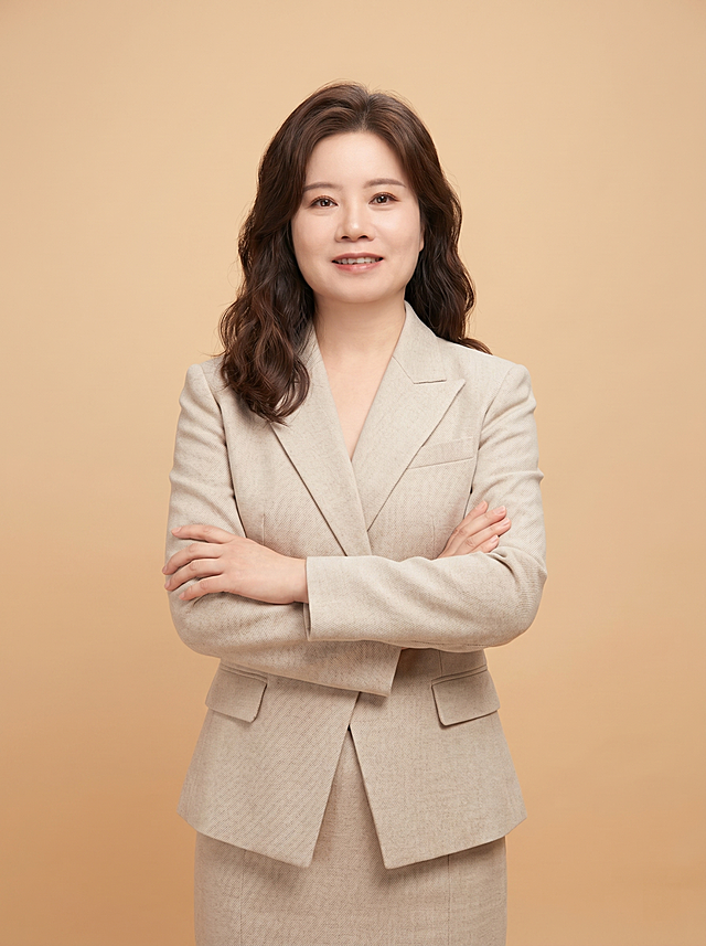 参考图中保留人物脸型、发型、年龄等特征生成金融法律从业者职场形象照,标准焦段50mm,f/4光圈(f/2.8-f/4区间),平视视角(镜头与人物视线齐平);人物占画面70%,人物居中(居中构图),位于画面中心,为视觉聚焦点,入境范围头部及以下至臀部下方10cm(半身像);左右留少量背景空间;头部与画面顶部保留5%空间;头部发型完整,头肩比例协调,人物结构准确;身穿米色精纺羊毛混纺西装外套,(平驳领设计,面料呈细腻微微哑光质感);下装同色同料半身裙;着装正式干练;严谨理性、值得信赖;身形匀称,姿态端正,身体挺拔。呈自然站姿，双手抱胸，肢体放松且自信;发质微微柔顺，微微蓬松。简约微微暖调暖灰米杏纯色背景(呈现均匀柔和质感),背景光线均匀柔和,强化色彩对比的高级感,极简干净;画质超高分辨率、细节清晰,现代精致感风格;肤质精细化处理;眼神直视镜头,目光坚定且澄澈,表情自然。嘴角上扬;清透自然裸妆(轻薄底妆+淡唇妆)妆感轻薄轻透裸肌感,通透感肤质,肤质自然细腻,肤色健康均匀,雾面柔光裸肌感,保留真实皮肤细节｡正面柔光补光,4800K中性光,面部光比1:1.5,色调明亮｡中近景写实摄影,对焦清晰,高清保留皮肤细节。分辨率4K,去除人物项链配饰，去除人物之外的所有元素。严格按照图片内容生成。画面比例3:4。
