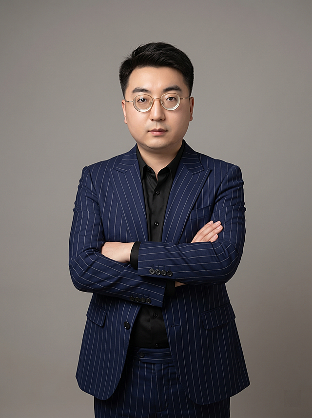 参考图中保留人物脸型、发型、年龄等特征生成职场形象照，标准焦段50mm，f/4光圈，平视视角，镜头与人物视线齐平；人物占画面70%，人物居中，居中构图，位于画面中心，为视觉聚焦点，入境范围头部及以下至臀部下方10cm，半身像；左右留少量背景空间；头部与画面顶部保留5%空间；头部发型完整，头肩比例协调，人物结构准确。发质微微柔顺，微微蓬松。身穿深蓝色细条纹精纺羊毛混纺西装外套，平驳领设计，面料呈细腻微微哑光质感；内搭黑色真丝缎面衬衫；下装深蓝色精纺羊毛混纺西裤；着装正式干练；专业稳重；身形匀称，姿态端正，身体挺拔。呈双手抱胸的自然站姿，肢体放松且自信；佩戴金属框眼镜。简约微微暖调中性灰色纯色背景，呈现均匀柔和质感，背景光线均匀柔和，强化色彩对比的高级感，极简干净。画质超高分辨率、细节清晰，现代精致感风格；肤质精细化处理；眼神直视镜头，目光坚定且澄澈，表情自然。肤色自然细腻。肤质自然细腻，肤色健康均匀，皮肤状态清爽干净，保留真实皮肤细节｡正面柔光补光，4800K中性光，面部光比1:1.5｡中近景写实摄影，对焦清晰，面部雾面柔光，轻透裸肌感，高清保留皮肤细节｡分辨率4K，去除人物之外的所有元素。严格按照图片内容生成，画面比例3:4。