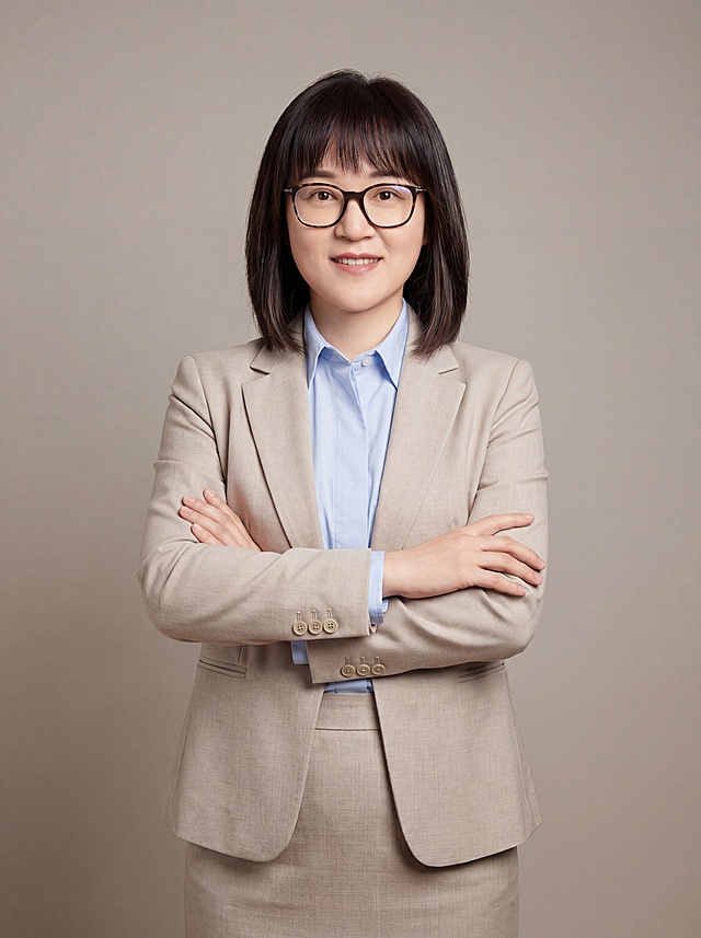 参考图中保留人物脸型、发型、年龄等特征生成职场形象照，标准焦段50mm，f/4光圈，平视视角，镜头与人物视线齐平；人物占画面70%，人物居中，居中构图，位于画面中心，为视觉聚焦点，入境范围头部及以下至臀部下方10cm，半身像；左右留少量背景空间；头部与画面顶部保留5%空间；头部发型完整，头肩比例协调，人物结构准确；发质微微柔顺，微微蓬松。身穿浅米色精纺羊毛混纺西装外套，平驳领设计，面料呈细腻微微哑光质感；内搭浅蓝色高支精梳棉衬衫；下装浅米色精纺羊毛混纺西装套裙；着装正式干练；专业亲切；身形匀称，姿态端正，身体挺拔。呈自然站姿，双手抱胸，肢体放松且自信；佩戴眼镜；简约暖灰色米杏纯色背景，呈现均匀柔和质感，背景光线均匀柔和，强化色彩对比的高级感，极简干净；画质超高分辨率、细节清晰，现代精致感风格；肤质精细化处理；眼神直视镜头，目光坚定且澄澈，表情自然。嘴角上扬；清透自然裸妆，轻薄底妆+淡唇妆，妆感轻薄轻透裸肌感，通透感肤质，肤质自然细腻，肤色健康均匀，雾面柔光裸肌感，保留真实皮肤细节。正面柔光补光，4800K中性光，面部光比1:1.5，色调明亮。中近景写实摄影，对焦清晰，高清保留皮肤细节；分辨率4K，去除人物之外的所有元素。严格按照图片内容生成，生成2寸形象照，画面比例3:4