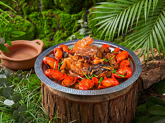 小龙虾焖鸡菜品，严格参照上传产品图（https://gdesign-dam-hz.dancf.com/36668346314905689/39c342f43d0346029615b7a904062b13.png），完整保留所有可见组件、部件、轮廓形状、结构、比例、材质质感、颜色与上传图完全一致。
户外自然绿植场景，贴近自然的清新绿色调。
产品放置在粗木原木承载面上，侧前方柔和自然光（还原参考图光影方向），产品受光面泛起清新绿色、暗部映射深棕木质光晕，与环境物理融合。
居中特写构图，微侧25°视角，自然静态展示。
全量换款清单：①承载木段：参考图横放粗木 → 生成图竖切粗木桩（同为实木，保留承载功能，换摆放形态）；②背景绿植：参考图大面积苔藓背景 → 生成图混合青草与尤加利叶背景（同为绿植，换具体植物种类）；③左侧奶壶：参考图白色陶瓷奶壶 → 生成图浅陶土小陶罐（同为盛器，换材质造型）；④右上角龟背竹：参考图大叶片龟背竹 → 生成图散尾葵叶片（同为热带绿植，换植物种类）；
消除品牌文字。4K分辨率，严格锁定宽高比为 4:3。