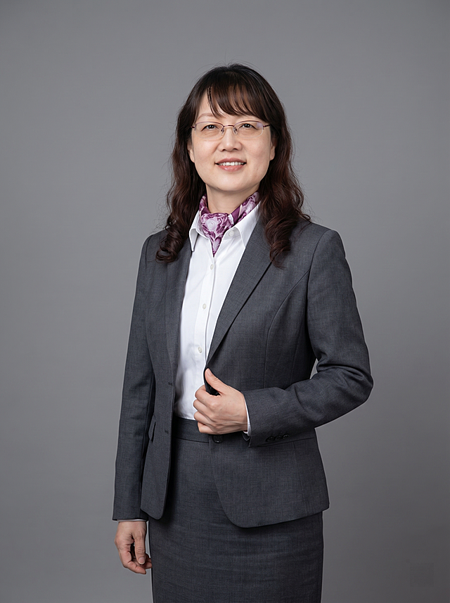 参考图中保留人物脸型、发型、年龄等特征生成职场形象照,标准焦段50mm,f/4光圈(f/2.8-f/4区间),平视视角(镜头与人物视线齐平);人物占画面70%,人物居中(居中构图),位于画面中心,为视觉聚焦点,入境范围头部及以下至臀部下方10cm(半身像);左右留少量背景空间;头部与画面顶部保留5%空间;头部发型完整,头肩比例协调,人物结构准确;发质微微柔顺，微微蓬松。身穿深灰色精纺羊毛混纺西装外套,(平驳领设计,面料呈细腻微微哑光质感);内搭白色高支精梳棉衬衫;下装深灰色精纺羊毛混纺西装套裙;着装正式干练;专业稳重;身形匀称,姿态端正,身体挺拔。呈自然站姿，右手轻握西装前襟，肢体放松且自信;佩戴精致配饰，保留眼镜和丝巾特征;简约微微中度灰色纯色背景(呈现均匀柔和质感),背景光线均匀柔和,强化色彩对比的高级感。极简干净;画质超高分辨率、细节清晰,现代精致感风格;肤质精细化处理;眼神直视镜头,目光坚定且澄澈,表情自然。嘴角上扬;清透自然裸妆(轻薄底妆+淡唇妆)妆感轻薄轻透裸肌感,通透感肤质,肤质自然细腻,肤色健康均匀,雾面柔光裸肌感,保留真实皮肤细节｡正面柔光补光,4800K中性光,面部光比1:1.5,色调明亮｡中近景写实摄影,对焦清晰,高清保留皮肤细节｡分辨率4K,去除人物之外的所有元素。严格按照图片内容生成。画面比例3:4