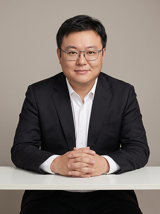 参考图中严格保留人物脸型、五官比例、神态与发型特征生成职场形象照,标准焦段50mm,f/4光圈(f/2.8-f/4区间),平视视角(镜头与人物视线齐平);人物占画面70%,人物居中(居中构图),位于画面中心,为视觉聚焦点,入境范围头部及以下至臀部下方10cm(半身像);左右留少量背景空间;头部与画面顶部保留5%空间;头部发型完整,头肩比例协调,人物结构准确;发质微微柔顺，微微蓬松。身穿深黑色精纺羊毛混纺西装外套,(平驳领设计,面料呈细腻微微哑光质感);内搭白色高支精梳棉衬衫;下装深黑色精纺羊毛混纺西裤;着装正式干练;专业稳重,值得信赖;身形匀称,姿态端正,身体挺拔。呈自然坐姿，双手交叠置于桌面边缘（桌面高度约80cm），手轻放于桌面边缘，肢体放松且自信;佩戴眼镜，保留眼镜特征;简约微微暖调暖灰/米杏纯色背景(呈现均匀柔和质感),背景光线均匀柔和,强化色彩对比的高级感,极简干净;画质超高分辨率、细节清晰,现代精致感风格;肤质精细化处理;眼神直视镜头,目光坚定且澄澈,表情自然。嘴角上扬;肤质自然细腻,肤色健康均匀,皮肤状态清爽干净,保留真实皮肤细节｡正面柔光补光,4800K中性光,面部光比1:1.5｡中近景写实摄影,对焦清晰,面部雾面柔光,轻透裸肌感,高清保留皮肤细节｡分辨率4K,去除人物之外的所有元素。严格按照图片内容生成。画面比例3:4