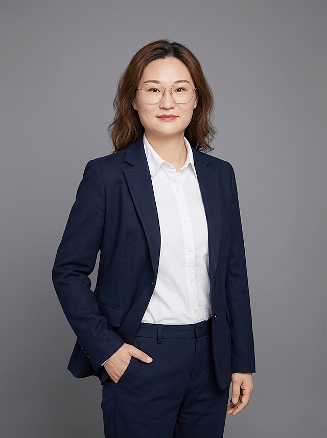 参考图中保留人物脸型、发型、年龄等特征生成教师职场形象照,标准焦段50mm,f/4光圈(f/2.8-f/4区间),平视视角(镜头与人物视线齐平);人物占画面70%,人物居中(居中构图),位于画面中心,为视觉聚焦点,入境范围头部及以下至臀部下方10cm(半身像);左右留少量背景空间;头部与画面顶部保留5%空间;头部发型完整,头肩比例协调,人物结构准确;身穿复古休闲藏青色精纺羊毛西装外套;内搭白色棉质衬衫;下装藏青色直筒休闲裤;着装正式干练;博学温润,知性内敛;身形匀称,姿态端正,身体挺拔。呈站姿，单手插兜的自然姿态，肢体放松且自信;佩戴透明细框眼镜;发质微微柔顺，微微蓬松。简约微微中度灰色纯色背景(呈现均匀柔和质感),背景光线均匀柔和,强化色彩对比的高级感。极简干净;画质超高分辨率、细节清晰,现代精致感风格;肤质精细化处理;眼神直视镜头,目光坚定且澄澈,表情自然。嘴角上扬;清透自然裸妆(轻薄底妆+淡唇妆)妆感轻薄轻透裸肌感,通透感肤质,肤质自然细腻,肤色健康均匀,雾面柔光裸肌感,保留真实皮肤细节｡正面柔光补光,4800K中性光,面部光比1:1.5,色调明亮｡中近景写实摄影,对焦清晰,高清保留皮肤细节｡分辨率4K,去除人物之外的所有元素,去除蓝色挂绳配饰。严格按照图片内容生成。画面比例3:4