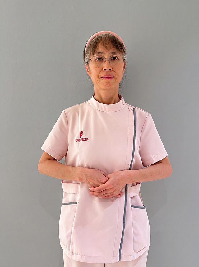 参考图中保留人物脸型、发型、年龄等特征生成医护职业形象照,标准焦段50mm,f/4光圈(f/2.8-f/4区间),平视视角(镜头与人物视线齐平);人物占画面70%,人物居中(居中构图),位于画面中心,为视觉聚焦点,入境范围头部及以下至臀部下方10cm(半身像);左右留少量背景空间;头部与画面顶部保留5%空间;头部发型完整,头肩比例协调,人物结构准确;身穿粉色医护工作服，保留灰色镶边和红色刺绣logo，面料干净整洁;下装同色医护裤;着装正式干练;专业可靠，温和严谨;身形匀称,姿态端正,身体挺拔。呈站姿，双手交叠于腹前的自然姿态，肢体放松且自信;保留发箍、眼镜配饰;发质微微柔顺，微微蓬松。简约微微中度灰色纯色背景(呈现均匀柔和质感),背景光线均匀柔和,强化色彩对比的高级感。极简干净;画质超高分辨率、细节清晰,现代精致感风格;肤质精细化处理;眼神直视镜头,目光坚定且澄澈,表情自然。嘴角上扬;清透自然裸妆(轻薄底妆+淡唇妆)妆感轻薄轻透裸肌感,通透感肤质,肤质自然细腻,肤色健康均匀,雾面柔光裸肌感,保留真实皮肤细节｡正面柔光补光,4800K中性光,面部光比1:1.5,色调明亮｡中近景写实摄影,对焦清晰,高清保留皮肤细节｡分辨率4K,去除人物之外的所有元素。严格按照图片内容生成，画面比例3:4。
