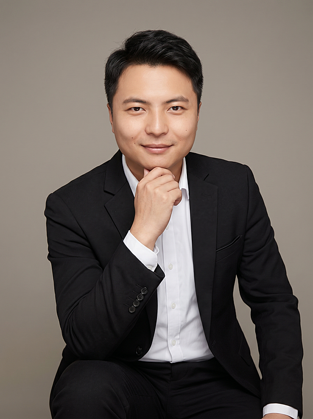 参考图中保留人物脸型、发型、年龄等特征生成互联网技术职场形象照，标准焦段50mm，f/4光圈(f/2.8-f/4区间)，平视视角(镜头与人物视线齐平)；人物占画面70%，人物居中(居中构图)，位于画面中心，为视觉聚焦点，入境范围头部及以下至臀部下方10cm(半身像)；左右留少量背景空间；头部与画面顶部保留5%空间；头部发型完整，头肩比例协调，人物结构准确；发质微微柔顺，微微蓬松。身穿深黑色精纺羊毛混纺西装外套，(平驳领设计，面料呈细腻微微哑光质感)；内搭白色高支精梳棉衬衫；下装深黑色精纺羊毛混纺西裤；着装正式干练；创新务实、年轻干练、沉稳高效；身形匀称，姿态端正，身体挺拔。呈坐姿，手扶下巴的自然姿态，肢体放松且自信；简约微微暖调中性灰纯色背景(呈现均匀柔和质感)，背景光线均匀柔和，强化色彩对比的高级感，极简干净；画质超高分辨率、细节清晰，现代精致感风格；肤质精细化处理；眼神直视镜头，目光坚定且澄澈，表情自然。嘴角上扬；肤质自然细腻，肤色健康均匀，皮肤状态清爽干净，保留真实皮肤细节。正面柔光补光，4800K中性光，面部光比1:1.5。中近景写实摄影，对焦清晰，面部雾面柔光，轻透裸肌感，高清保留皮肤细节；分辨率4K，去除人物配饰，去除人物之外的所有元素。严格按照图片内容生成。画面比例3:4