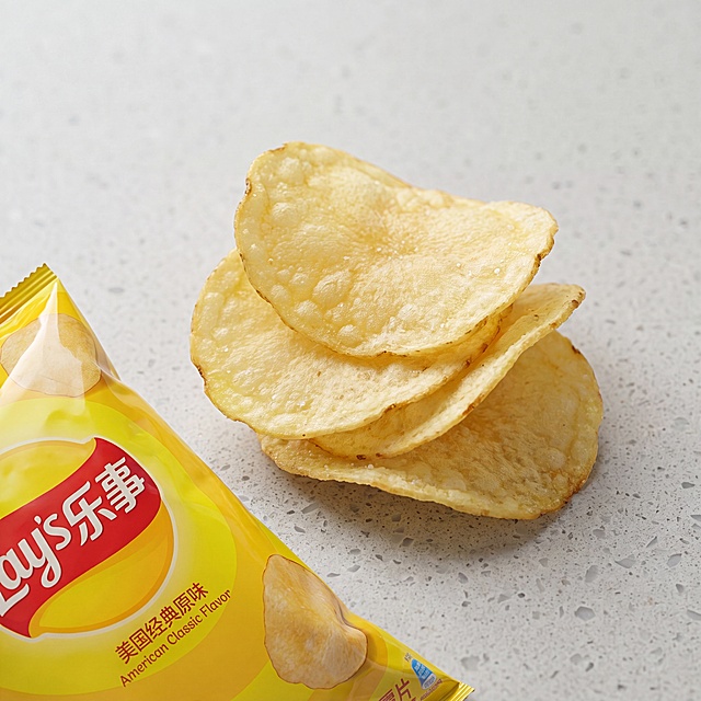 1:1 正方形微距食材特写，薯片原料材质细节展示图，严格还原上传的产品图，包括包装设计、颜色、LOGO 位置、文字内容、图案元素等所有细节，产品外观必须与参考图完全一致，不得改变包装设计、配色方案或品牌元素，保持产品的真实材质质感（高光塑料包装 + 薯片哑光薯肉肌理）。画面左下角只露出小部分乐事原装黄色包装袋边角，保留局部 LOGO 与口味文字印刷细节，画面主体为三片错落堆叠的原味薯片，微距近距离拍摄，完整捕捉薄切工艺带来的自然弧度、烘烤形成的细密气孔、马铃薯原生浅黄肉质纹路、表层细微调味盐粒颗粒。承载台面为浅灰色哑光水磨石材质，顶侧均匀漫射柔光，薯片缝隙形成自然浅淡软阴影，8K 超高清美食写实画质，色彩贴合食材原生本色，无过度增亮、夸张增色。画面聚焦薯片原料与制作材质卖点，直观体现鲜薯原料、薄切酥脆产品优势，画面干净极简无多余摆件，适配产品原料卖点详情配图，纯实物实拍。负面词：薯片人工染色过重、包装颜色篡改、LOGO 残缺不全、表面厚重浮油、软烂受潮薯片、霉斑黑点、动漫插画、3D 渲染建模、水印文字、画面糊片、强光刺眼爆亮、全黑死角、多余酱料辅料、畸形不规则薯片、合成拼接画面、荧光怪异色、密集颗粒噪点、多余果蔬摆件、台面划痕破损、印刷错乱文字、哑光包装（原高光）、手绘纹理、特效滤镜、杂乱碎屑、虚假膨化体积、冷蓝偏色、多余包装碎片