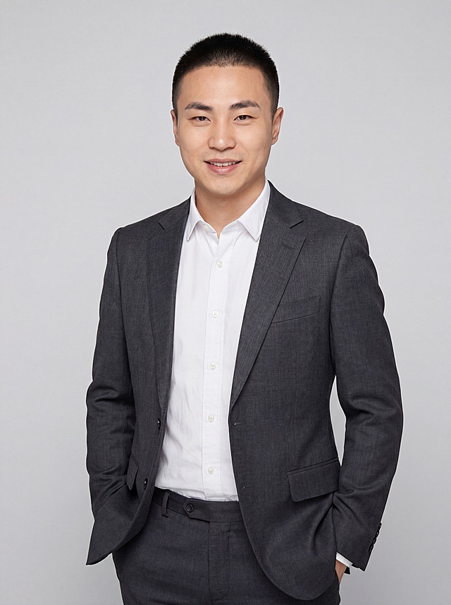 参考图中保留人物脸型、发型、年龄等特征生成教师/学术职场形象照,标准焦段50mm,f/4光圈(f/2.8-f/4区间),平视视角(镜头与人物视线齐平);人物占画面70%,人物居中(居中构图),位于画面中心,为视觉聚焦点,入境范围头部及以下至臀部下方10cm(半身像);左右留少量背景空间;头部与画面顶部保留5%空间;头部发型完整,头肩比例协调,人物结构准确;发质微微柔顺，微微蓬松。身穿深灰色精纺羊毛混纺西装外套,(平驳领设计,面料呈细腻微微哑光质感);内搭白色高支精梳棉衬衫;下装深灰色精纺羊毛混纺西裤;着装正式干练;博学温润,学者风范;身形匀称,姿态端正,身体挺拔。呈站姿，双手插兜的自然姿态，肢体放松且自信;简约浅灰色纯色背景(呈现均匀柔和质感),背景光线均匀柔和,强化色彩对比的高级感。极简干净;画质超高分辨率、细节清晰,现代精致感风格;肤质精细化处理;眼神直视镜头,目光坚定且澄澈,表情自然。嘴角上扬;肤质自然细腻,肤色健康均匀,皮肤状态清爽干净,保留真实皮肤细节｡正面柔光补光,4800K中性光,面部光比1:1.5｡中近景写实摄影,对焦清晰,面部雾面柔光,轻透裸肌感,高清保留皮肤细节｡分辨率4K,去除人物之外的所有元素。严格按照图片内容生成。画面比例3:4
