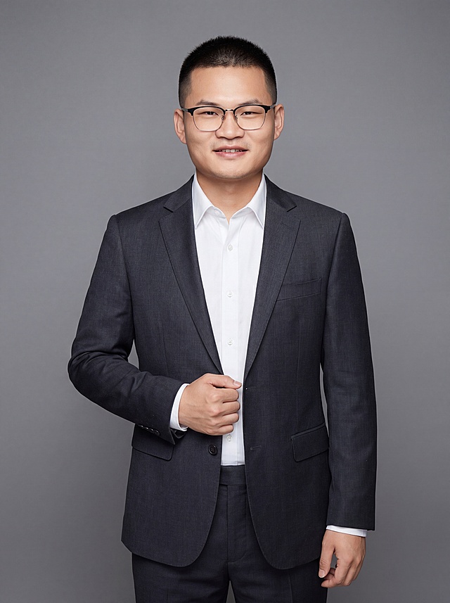 参考图中保留人物脸型、发型、年龄等特征生成职场形象照,标准焦段50mm,f/4光圈(f/2.8-f/4区间),平视视角(镜头与人物视线齐平);人物占画面70%,人物居中(居中构图),位于画面中心,为视觉聚焦点,入境范围头部及以下至臀部下方10cm(半身像);左右留少量背景空间;头部与画面顶部保留5%空间;头部发型完整,头肩比例协调,人物结构准确;发质微微柔顺，微微蓬松。身穿深炭灰色精纺羊毛混纺西装外套,(平驳领设计,面料呈细腻微微哑光质感);内搭白色高支精梳棉衬衫;下装深炭灰色精纺羊毛混纺西裤;着装正式干练;专业稳重;身形匀称,姿态端正,身体挺拔。呈自然站姿，右手轻握西装前襟，肢体放松且自信;佩戴黑框眼镜，保留配饰特征;简约微微中度灰色纯色背景(呈现均匀柔和质感),背景光线均匀柔和,强化色彩对比的高级感。极简干净;画质超高分辨率、细节清晰,现代精致感风格;肤质精细化处理;眼神直视镜头,目光坚定且澄澈,表情自然。嘴角上扬;肤质自然细腻,肤色健康均匀,皮肤状态清爽干净,保留真实皮肤细节｡正面柔光补光,4800K中性光,面部光比1:1.5｡中近景写实摄影,对焦清晰,面部雾面柔光,轻透裸肌感,高清保留皮肤细节｡分辨率4K,去除人物之外的所有元素。严格按照图片内容生成。画面比例3:4