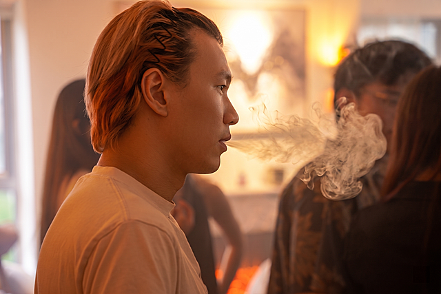 使用相同方式精修图中前景人物：提升面部精致度（皮肤细腻、轮廓清晰、眼神自然有神），增强光影层次与肤色通透感，保持真实写实风格；烟雾效果保留朦胧美感但不遮挡主体面部细节; 背景适度虚化以突出人物侧脸轮廓，保留原暖色调氛围