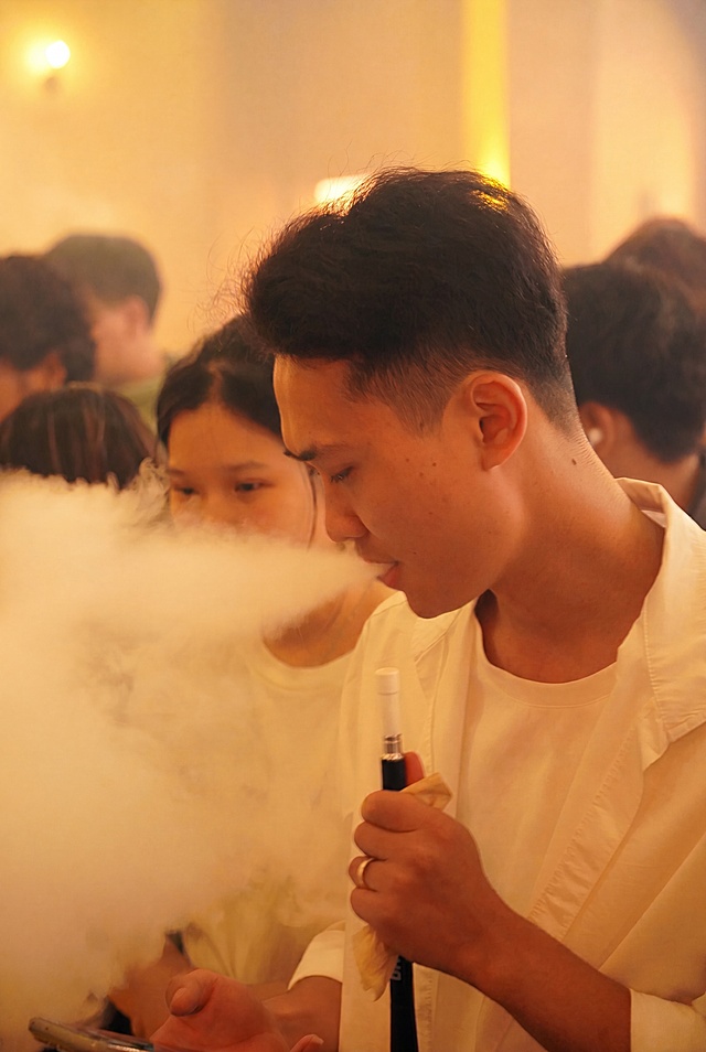 图生图：仅去除画面中的模糊与烟雾遮挡感，提升通透度与清晰度；严格保持原图竖屏比例，不改为横屏；不改变任何细节——包括前景人物发型（黑色短发）、衣着（白色衬衫）、手持电子烟动作、呼出烟雾形态，以及背景中人物轮廓与暖黄色调环境; 4K超清画质，真实摄影风格，自然光线，细节锐利
