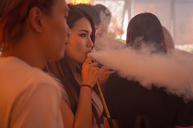 使用相同方式精修图中前景人物（正在吸电子烟的女性）：提升面部精致度（皮肤细腻光滑、眼神专注有神、轮廓清晰立体），增强光影层次与肤色通透感，保持真实写实风格；烟雾效果保留自然流动感与朦胧氛围，但不遮挡面部关键细节；背景适度虚化以突出主体，维持原暖色调光影氛围