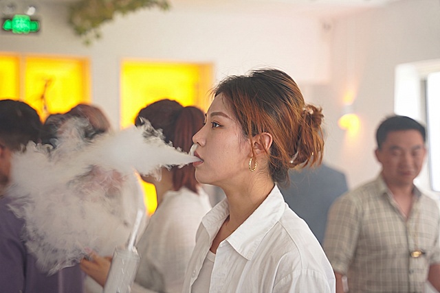 仅进行极轻微的画质增强：提升前景女性面部清晰度、皮肤质感与眼神光，优化烟雾的层次感与通透度，保持真实写实风格；严格不改变发型（发色、丸子头造型、发饰）、服饰（白衬衫细节）、耳环、姿态、手持动作、背景人物虚化程度、暖黄色灯光氛围及构图——所有元素位置、形态、比例均与原图一致
