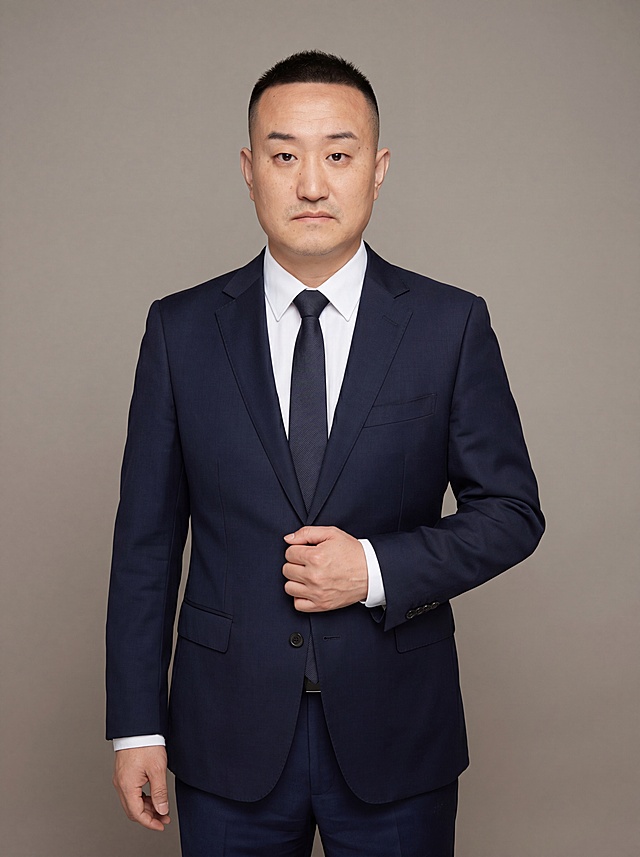 参考图中保留人物脸型、发型、年龄等特征生成金融法律从业者职场形象照，标准焦段50mm，f/4光圈，平视视角，镜头与人物视线齐平；人物占画面70%，人物居中，居中构图，位于画面中心，为视觉聚焦点，入境范围头部及以下至臀部下方10cm，半身像；左右留少量背景空间；头部与画面顶部保留5%空间；头部发型完整，头肩比例协调，人物结构准确；发质微微柔顺，微微蓬松。身穿藏青色精纺羊毛混纺正装西装外套，平驳领设计，面料呈细腻微微哑光质感；内搭白色高支精梳棉素色长袖衬衫；下装藏青色精纺羊毛混纺直筒西装裤；佩戴低调素色领带；着装正式干练；严谨理性，逻辑清晰，不怒自威，公信力强；身形匀称，姿态端正，身体挺拔。呈站姿右手轻握西装前襟的自然姿态，肢体放松且自信；简约微微暖调中性灰色纯色背景，呈现均匀柔和质感，背景光线均匀柔和，强化色彩对比的高级感，极简干净；画质超高分辨率、细节清晰，现代精致感风格；肤质精细化处理；眼神直视镜头，目光坚定且澄澈，表情自然；去除人物原衬衫，替换为指定西装，去除配饰。肤质自然细腻，肤色健康均匀，皮肤状态清爽干净，保留真实皮肤细节｡正面柔光补光，4800K中性光，面部光比1:1.5｡中近景写实摄影，对焦清晰，面部雾面柔光，轻透裸肌感，高清保留皮肤细节｡分辨率4K，去除人物之外的所有元素。严格按照图片内容生成。画面比例3:4