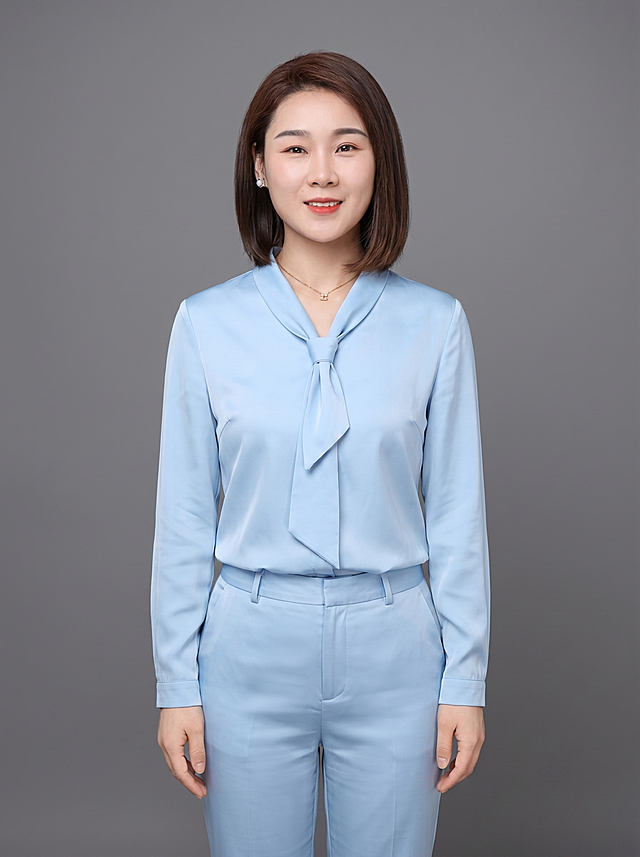 参考图中保留人物脸型、发型、年龄等特征生成职场形象照，标准焦段50mm，f/4光圈，平视视角（镜头与人物视线齐平）；人物占画面，人物居中（居中构图），位于画面中心，为视觉聚焦点，含腿的全身照，展现人物完整身形；头部与画面顶部保留5%空间；头部发型完整，头身比例协调，人物结构准确；身穿浅蓝色真丝缎面衬衫（翻领系带设计，面料呈细腻微微哑光质感）；下装浅蓝色同面料西装裤；着装正式干练；专业稳重；身形匀称，姿态端正，身体挺拔。呈自然站姿，双手自然下垂，肢体放松且自信；发质微微柔顺，微微蓬松；佩戴精致配饰（耳环和项链）；简约微微中度灰色纯色背景（呈现均匀柔和质感），背景光线均匀柔和，强化色彩对比的高级感。极简干净；画质超高分辨率、细节清晰，现代精致感风格；肤质精细化处理；眼神直视镜头，目光坚定且澄澈，表情自然，嘴角上扬；清透自然裸妆（轻薄底妆+淡唇妆）妆感轻薄轻透裸肌感，通透感肤质，肤质自然细腻，肤色健康均匀，雾面柔光裸肌感，保留真实皮肤细节｡正面柔光补光，4800K中性光，面部光比1:1.5，色调明亮｡中近景写实摄影，对焦清晰，高清保留皮肤细节；分辨率4K，去除人物之外的所有元素，严格按照图片内容生成；画面比例3:4
