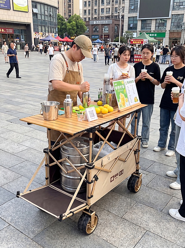 使用参考图中的卡其色露营推车，保留后开拖斗设计搭配折叠桌面，制作户外售卖手打柠檬茶场景，摊主正在制作柠檬茶，有顾客在旁边等待，背景为城市广场，推车上的折叠桌面上摆放着制作柠檬茶的装备，推车内放置一个不锈钢铁桶，8K超清，严格保留产品的设计和外观，图片整体比例为3:4