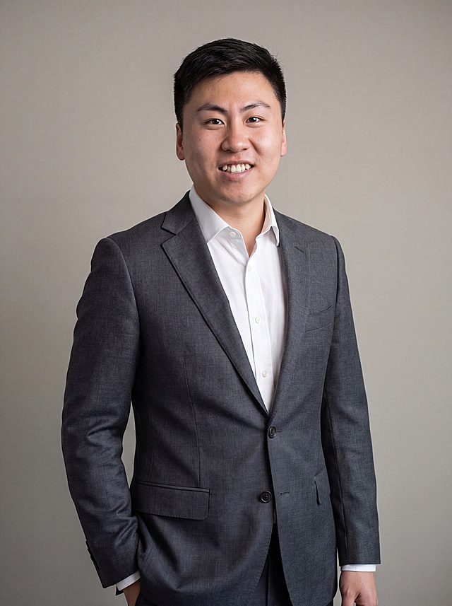 参考图中保留人物脸型、发型、年龄等特征生成职场形象照，标准焦段50mm，f/4光圈，平视视角，镜头与人物视线齐平；人物占画面70%，人物居中，居中构图，位于画面中心，为视觉聚焦点，入境范围头部及以下至臀部下方10cm（半身像）；左右留少量背景空间；头部与画面顶部保留5%空间；头部发型完整，头肩比例协调，人物结构准确；发质微微柔顺，微微蓬松。身穿深灰色精纺羊毛混纺西装外套，平驳领设计，面料呈细腻微微哑光质感；内搭白色高支精梳棉衬衫；下装深灰色精纺羊毛混纺西裤；着装正式干练；专业稳重；身形匀称，姿态端正，身体挺拔。呈左手插兜的自然站姿，肢体放松且自信；简约微微暖调中性灰纯色背景，呈现均匀柔和质感，背景光线均匀柔和，强化色彩对比的高级感，极简干净；画质超高分辨率、细节清晰，现代精致感风格；肤质精细化处理；眼神直视镜头，目光坚定且澄澈，表情自然，嘴角上扬；肤质自然细腻，肤色健康均匀，皮肤状态清爽干净，保留真实皮肤细节。正面柔光补光，4800K中性光，面部光比1:1.5；中近景写实摄影，对焦清晰，面部雾面柔光，轻透裸肌感，高清保留皮肤细节；分辨率4K，去除人物之外的所有元素，严格按照图片内容生成，画面比例3:4