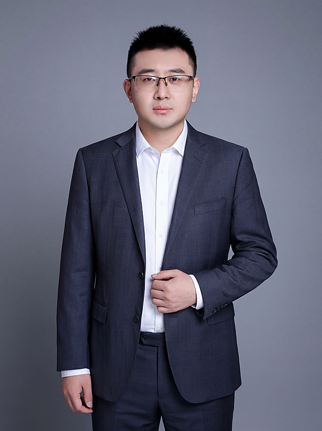 参考图中保留人物脸型、发型、年龄等特征生成金融商务职场形象照，标准焦段50mm，f/4光圈，平视视角（镜头与人物视线齐平）；人物占画面70%，人物居中，位于画面中心，为视觉聚焦点，入境范围头部及以下至臀部下方10cm（半身像）；左右留少量背景空间；头部与画面顶部保留5%空间；头部发型完整，头肩比例协调，人物结构准确；发质微微柔顺，微微蓬松。身穿深灰色精纺羊毛混纺纯色西装外套，平驳领设计，面料呈细腻微微哑光质感；内搭白色高支精梳棉衬衫；下装深灰色精纺羊毛混纺西裤；着装正式干练；权威沉稳、值得信赖；身形匀称，姿态端正，身体挺拔。呈站姿，右手轻握西装前襟的自然姿态，肢体放松且自信；佩戴黑框眼镜，保留配饰特征。简约冷灰纯色背景，呈现均匀柔和质感，背景光线均匀柔和，强化色彩对比的高级感，极简干净。画质超高分辨率、细节清晰，现代精致感风格；肤质精细化处理；眼神直视镜头，目光坚定且澄澈，表情自然。肤质自然细腻，肤色健康均匀，皮肤状态清爽干净，保留真实皮肤细节。正面柔光补光，4800K中性光，面部光比1:1.5。中近景写实摄影，对焦清晰，面部雾面柔光，轻透裸肌感，高清保留皮肤细节。分辨率4K，去除人物之外的所有元素。严格按照图片内容生成。画面比例3:4