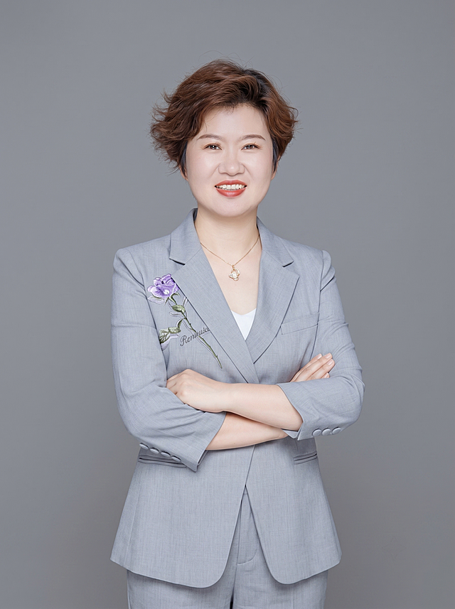 参考图中保留人物脸型、发型、年龄等特征生成眼科医生职业形象照,标准焦段50mm,f/4光圈(f/2.8-f/4区间),平视视角(镜头与人物视线齐平);人物占画面70%,人物居中(居中构图),位于画面中心,为视觉聚焦点,入境范围头部及以下至臀部下方10cm(半身像);左右留少量背景空间;头部与画面顶部保留5%空间;头部发型完整,头肩比例协调,人物结构准确;发质微微柔顺，微微蓬松。身穿浅灰色精纺羊毛混纺西装外套,(平驳领设计,面料呈细腻微微哑光质感)，衣服保留紫色花刺绣装饰;内搭白色打底;下装同浅灰色精纺羊毛混纺西装裤;着装正式干练;专业仁心、沉稳可靠;身形匀称,姿态端正,身体挺拔。呈自然站姿，双手抱胸，肢体放松且自信;佩戴精致项链配饰;简约微微中度灰色纯色背景(呈现均匀柔和质感),背景光线均匀柔和,强化色彩对比的高级感。极简干净;画质超高分辨率、细节清晰,现代精致感风格;肤质精细化处理;眼神直视镜头,目光坚定且澄澈,表情自然。嘴角上扬;清透自然裸妆(轻薄底妆+淡唇妆)妆感轻薄轻透裸肌感,通透感肤质,肤质自然细腻,肤色健康均匀,雾面柔光裸肌感,保留真实皮肤细节｡正面柔光补光,4800K中性光,面部光比1:1.5,色调明亮｡中近景写实摄影,对焦清晰,高清保留皮肤细节｡分辨率4K,去除人物之外的所有原有文字和装饰元素。严格按照图片内容生成。画面比例3:4