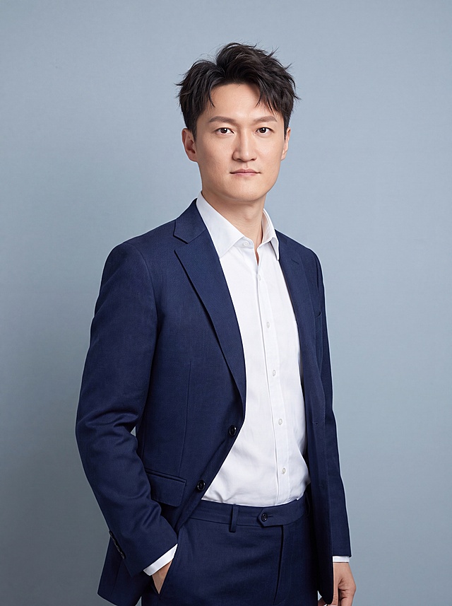 参考图中保留人物脸型特征，生成一张年轻的企业高管半身职业形象照，让人物显得更年轻，设计更时尚干练的发型，身穿藏青色定制精纺羊毛混纺西装外套（平驳领设计，面料呈细腻微微哑光质感），内搭白色高支精梳棉衬衫，下装同料藏青色精纺羊毛混纺西裤，去除所有配饰，着装正式干练，权威沉稳，格局开阔，从容果决，值得信赖；身形匀称，姿态端正，身体挺拔，呈站姿，单手插兜，肢体放松且自信；发质微微柔顺，微微蓬松，更显时尚，主体锐利。简约冷灰蓝纯色背景，呈现均匀柔和质感，背景光线均匀柔和，强化色彩对比的高级感，极简干净。画质超高分辨率、细节清晰，现代精致感风格；肤质精细化处理；眼神直视镜头，目光坚定且澄澈，表情自然。肤质自然细腻，肤色健康均匀，皮肤状态清爽干净，保留真实皮肤细节｡正面柔光补光，4800K中性光，面部光比1:1.5｡中近景写实摄影，对焦清晰，面部雾面柔光，轻透裸肌感，高清保留皮肤细节；参考图中保留人物脸型、发型基础特征，调整得更年轻，发质蓬松时尚；分辨率4K，去除人物之外的所有元素。严格按照图片内容生成。画面比例3:4