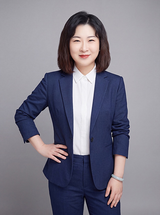 参考图中保留女性人物脸型、发型、年龄等脸部特征，生成教师职业形象照，标准焦段50mm，f/4光圈，平视视角，镜头与人物视线齐平，人物占画面70%，人物居中构图，位于画面中心，为视觉聚焦点，半身照，左右留少量背景空间，头部与画面顶部保留5%空间，头部发型完整，头肩比例协调，人物结构准确。身穿藏蓝色精纺羊毛混纺西装外套，平驳领设计，面料呈细腻微微哑光质感；内搭白色衬衫；下装藏蓝色精纺羊毛混纺西裤；着装正式干练，知性专业，身形匀称，姿态端正，身体挺拔。呈自然站姿，单手叉腰，肢体放松且自信；保留玉镯配饰；发梢微微修整，干练大气，主体锐利，背景光线均匀柔和，强化色彩对比的高级感。中性灰纯色背景，呈现均匀柔和质感，极简干净；画质超高分辨率，细节清晰，现代精致感风格；肤质精细化处理，眼神直视镜头，目光坚定且澄澈，表情自然，嘴角上扬；肤质自然细腻，肤色健康均匀，哑光肤质清爽干净，保留真实皮肤细节；杰作，最佳画质，超高细节，写实摄影，中近景人像，皮肤质感自然清晰，柔光，正面补光，画面干净清新，高清皮肤细节；面部光比1:1.5，4800K中性光，微微修饰脸型，肤质哑光细腻，面部雾面柔光，轻透裸肌感，真实毛孔纹理；分辨率4K，严格保留脸部特征，画面比例3:4