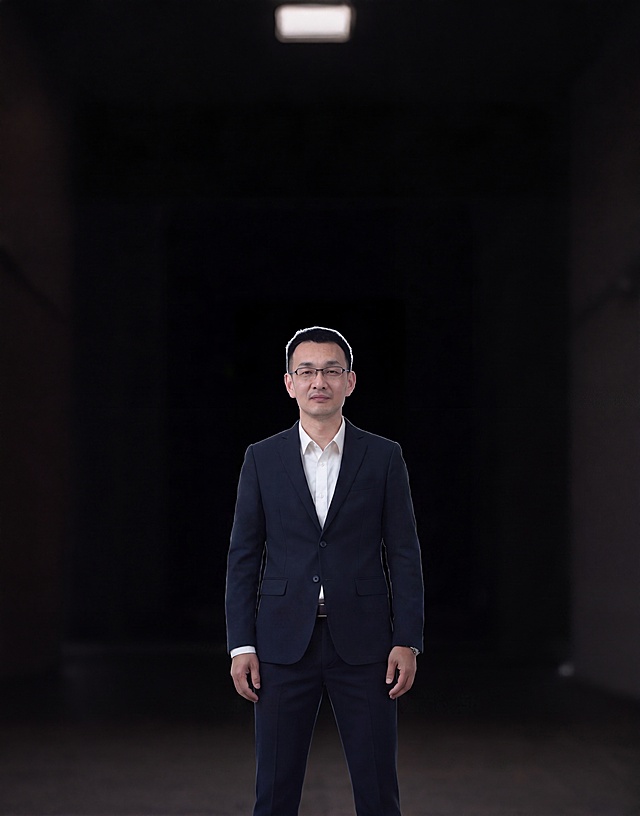 修改图中男士，将双手从口袋中拿出，自然垂放在身体两侧，保持男士原本穿着深蓝色西装白色衬衫，保持黑色背景走廊场景，整体姿势自然协调，不改变人物样貌和整体构图