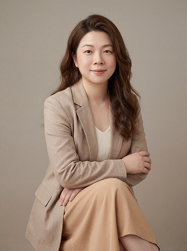参考图中保留人物脸型、发型、年龄等特征生成情感主播职场形象照,标准焦段50mm,f/4光圈(f/2.8-f/4区间),平视视角(镜头与人物视线齐平);人物占画面70%,人物居中(居中构图),位于画面中心,为视觉聚焦点,入境范围头部及以下至臀部下方10cm(半身像);左右留少量背景空间;头部与画面顶部保留5%空间;头部发型完整,头肩比例协调,人物结构准确;身穿莫兰迪暖米色棉麻西装外套,(平驳领设计,面料呈现自然肌理感);内搭米白色针织内搭;下装同料暖杏色垂感半裙;着装正式干练;温暖亲和、沉静治愈;身形匀称,姿态端正,身体挺拔。呈坐姿双手抱胸的自然姿态，肢体放松且自信;发质微微柔顺，微微蓬松。简约微微暖调暖灰米杏纯色背景(呈现均匀柔和质感),背景光线均匀柔和,强化色彩对比的高级感,极简干净;画质超高分辨率、细节清晰,现代精致感风格;肤质精细化处理;眼神直视镜头,目光坚定且澄澈,表情自然。嘴角上扬;清透自然裸妆(轻薄底妆+淡唇妆)妆感轻薄轻透裸肌感,通透感肤质,肤质自然细腻,肤色健康均匀,雾面柔光裸肌感,保留真实皮肤细节｡正面柔光补光,4800K中性光,面部光比1:1.5,色调明亮｡中近景写实摄影,对焦清晰,高清保留皮肤细节｡分辨率4K,去除人物之外的所有元素，去除项链和眼镜配饰。严格按照图片内容生成。画面比例3:4