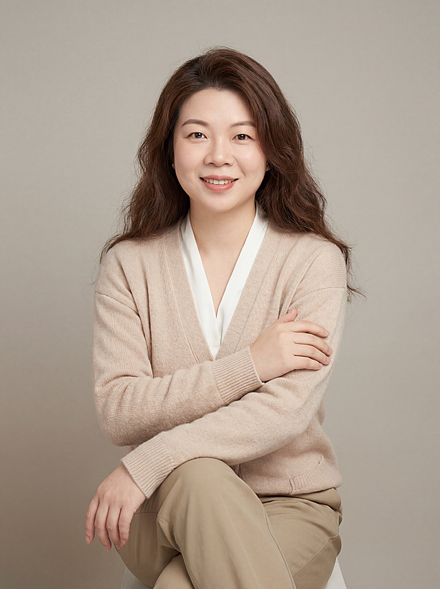 参考图中保留人物脸型、发型、年龄等特征生成情感主播职场形象照,标准焦段50mm,f/4光圈(f/2.8-f/4区间),平视视角(镜头与人物视线齐平);人物占画面70%,人物居中(居中构图),位于画面中心,为视觉聚焦点,入境范围头部及以下至臀部下方10cm(半身像);左右留少量背景空间;头部与画面顶部保留5%空间;头部发型完整,头肩比例协调,人物结构准确;身穿浅奶茶色针织开衫,(V领设计,面料软糯亲肤);内搭奶白色真丝衬衫;下装卡其色直筒休闲裤;着装柔和干练;温暖包容、耐心亲和;身形匀称,姿态端正,身体挺拔。呈坐姿，左手轻搭右手臂，双手抱胸自然姿态，肢体放松且自信;发质微微柔顺，微微蓬松。简约微微暖调暖灰米杏纯色背景(呈现均匀柔和质感),背景光线均匀柔和,强化色彩对比的高级感,极简干净;画质超高分辨率、细节清晰,现代精致感风格;肤质精细化处理;眼神直视镜头,目光坚定且澄澈,表情自然。嘴角上扬;清透自然裸妆(轻薄底妆+淡唇妆)妆感轻薄轻透裸肌感,通透感肤质,肤质自然细腻,肤色健康均匀,雾面柔光裸肌感,保留真实皮肤细节｡正面柔光补光,4800K中性光,面部光比1:1.5,色调明亮｡中近景写实摄影,对焦清晰,高清保留皮肤细节｡分辨率4K,去除人物之外的所有元素，去除项链和眼镜配饰。严格按照图片内容生成。画面比例3:4