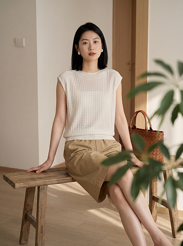 服装细节严格不动：保持原图所有服装版型、颜色，圆领设计领口贴合颈部，扁平方格编织肌理，每个小方块上有一条斜向纹理，面料纹理细节严格参考图2，面料极轻薄轻透，微透内衣透肤色，呈现细腻通透轻盈质感，下摆细密罗纹收口，材质莱赛尔纤维72.1%醋酯纤维27.9%，天丝醋酸混纺质感，丝滑垂坠，卡其色高腰中裤，棕色编织手提包，白色圆形耳饰全部保留不变。全身照，完整展示从头到脚。仅改变模特姿势：年轻东亚女性模特，黑色齐肩短发，身材修长，国际高端女装品牌夏日室内大片，日系知性优雅，法式极简高级，Slow Living生活方式摄影。姿势4：模特坐在木质长条凳上，双腿伸直微微交叉，双手放在身侧撑在凳子上，编织包放在右腿边，舒展随性。拍摄场景不变：室内浅色墙面，门框结构，绿植叶片前景虚化，自然侧光形成柔和明暗对比，光线柔和温暖，低饱和大地色系，知性优雅氛围。生活方式室内构图，突出质感从容与知性气质，中画幅摄影质感，真实面料纹理与轻薄微透方格细节，Kodak Portra胶片色彩，暖调低饱和，超写实，8K