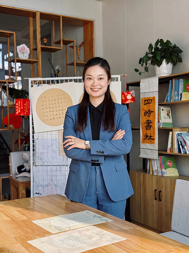 修改姿势，保持内搭黑色衬衣外穿蓝色西装的穿搭不变，保持原自然微笑表情和配饰不变，保持原办公室背景不变；改为双手交叉抱在胸前，身体向前倾斜，整体姿势自然，画面协调，保持专业职业形象，画质清晰。