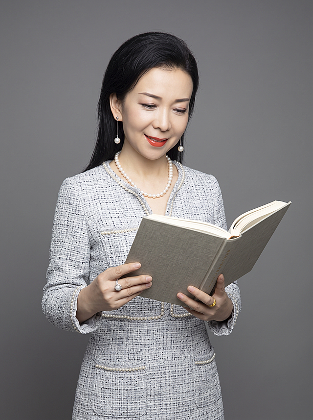 参考图生成，保留原人物长相五官特征不变，半身形象照，摄影棚级，背景换成干净的中性灰色纯色背景，穿搭为浅灰色小香风连衣裙，手持书本阅读姿势，保持自然真实感，不改变长相，专业打光，画质高清，半身形象照，分辨率4K