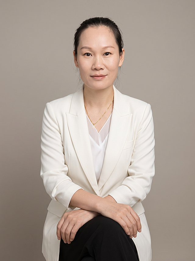 参考图中保留人物脸型、发型、年龄等特征生成职场形象照，标准焦段50mm，f/4光圈(f/2.8-f/4区间)，平视视角(镜头与人物视线齐平)；人物占画面70%，人物居中(居中构图)，位于画面中心，为视觉聚焦点，入境范围头部及以下至臀部下方10cm(半身像)；左右留少量背景空间；头部与画面顶部保留5%空间；头部发型完整，头肩比例协调，人物结构准确；发质微微柔顺，微微蓬松；身穿白色精纺羊毛混纺西装外套，平驳领设计，面料呈细腻微微哑光质感；内搭白色V领真丝拼接内搭；下装黑色精纺西装西裤；着装正式干练；专业稳重，亲和得体；身形匀称，姿态端正，身体挺拔。呈自然坐姿，双手交叠放在腿上，肢体放松且自信；佩戴精致金项链；简约微微暖调暖灰米杏纯色背景，呈现均匀柔和质感，背景光线均匀柔和，强化色彩对比的高级感，极简干净；画质超高分辨率、细节清晰，现代精致感风格；肤质精细化处理；眼神直视镜头，目光坚定且澄澈，表情自然。嘴角上扬；清透自然裸妆，轻薄底妆+淡唇妆，妆感轻薄轻透裸肌感，通透感肤质，肤质自然细腻，肤色健康均匀，雾面柔光裸肌感，保留真实皮肤细节；正面柔光补光，4800K中性光，面部光比1:1.5，色调明亮；中近景写实摄影，对焦清晰，高清保留皮肤细节；分辨率4K，去除人物之外的其他多余元素，严格按照参考图片人物特征生成，画面比例3:4