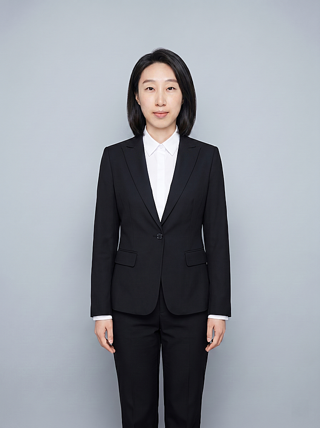 参考图保留人物脸型五官和所有服装不变，只修改发型，将发型调整得更长一些，长度达到齐肩，保持发型干练整齐，适配公务员职业形象，保持整体风格正式稳重，保持全身站姿不变，冷灰背景不变，画质保持超高分辨率细节清晰，清透自然裸肌感，保留真实皮肤细节，正面柔光补光，4800K中性光