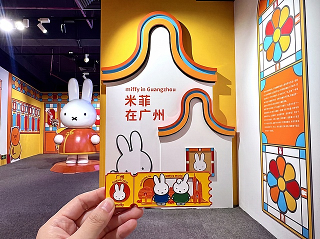 生成一张人手拿着广州米菲主题冰箱贴的图片，背景替换为第二张图的米菲展览室内场景