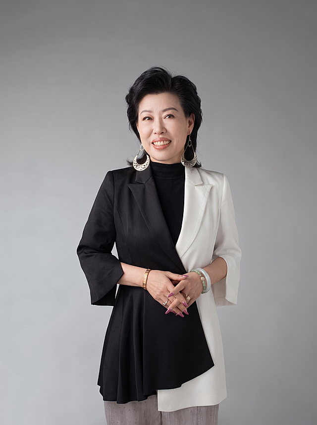 参考图中人物脸型、发型、年龄特征保持不变，生成高清专业企业高管形象照。标准焦段50mm，f/4光圈，平视视角，人物居中占画面70%，半身照，头部与顶部留5%空间，头肩比例协调。身穿黑白拼色精纺西装外套，左半侧黑色右半侧白色，七分袖设计，面料呈细腻哑光质感；内搭黑色高领打底；下装与上装风格协调；着装正式干练，气场强大，身形匀称姿态端正挺拔。呈站姿，双手交叠于腹前，肢体放松自信。佩戴耳环、手镯、戒指等精致配饰。发梢干练大气，主体锐利。中性灰纯色背景，均匀柔和质感，极简干净。画质超高分辨率、细节清晰，现代精致感风格。眼神直视镜头，目光坚定，表情自然微笑。肤质自然细腻，肤色健康均匀，哑光肤质清爽干净，保留真实皮肤细节。杰作，最佳画质，超高细节，写实摄影，中近景人像，皮肤质感自然清晰，柔光，正面补光，画面干净清新，高清皮肤细节。面部光比1:1.5，4800K中性光，微微修饰脸型，肤质哑光细腻，面部雾面柔光，轻透裸肌感，真实毛孔纹理。画面比例3:4。