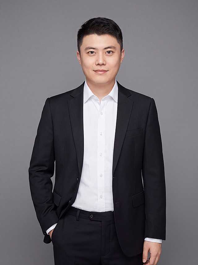 参考图中保留人物五官、脸型、发型、年龄等特征生成职场半身形象照，清晰度增强，标准焦段50mm，f/4光圈，平视视角，镜头与人物视线齐平；人物占画面70%，人物居中构图，位于画面中心，为视觉聚焦点，入境范围头部及以下至臀部下方10cm（半身像）；左右留少量背景空间；头部与画面顶部保留5%空间；头部发型完整，头肩比例协调，人物结构准确；身穿深黑色精纺羊毛混纺西装外套，平驳领设计，面料呈细腻微微哑光质感；内搭白色高支精梳棉衬衫；下装深黑色精纺羊毛混纺西裤；着装正式干练；专业稳重；身形匀称，姿态端正，身体挺拔。呈自然站姿，左手插兜的自然姿态，肢体放松且自信；发质微微柔顺，微微蓬松。简约微微中度灰色纯色背景，呈现均匀柔和质感，背景光线均匀柔和，强化色彩对比的高级感，极简干净；画质超高分辨率、细节清晰，现代精致感风格；肤质精细化处理；眼神直视镜头，目光坚定且澄澈，表情自然，嘴角上扬；肤质自然细腻，肤色健康均匀，皮肤状态清爽干净，保留真实皮肤细节。正面柔光补光，4800K中性光，面部光比1:1.5。中近景写实摄影，对焦清晰，面部雾面柔光，轻透裸肌感，高清保留皮肤细节。分辨率4K，去除人物之外的所有元素，严格按照图片内容生成，画面比例3:4