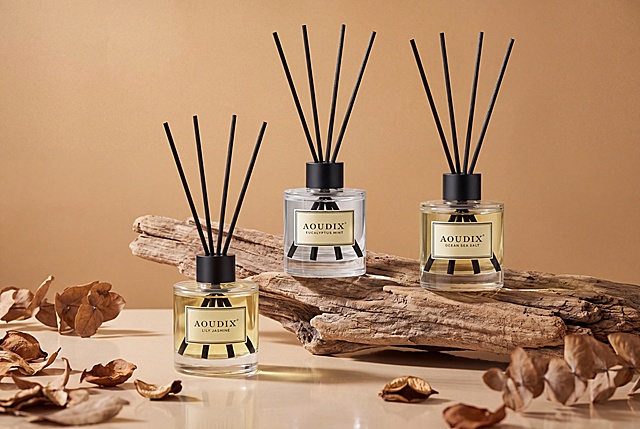 3:2横版构图，高低错落双主体：三瓶香薰错落排布，左侧靠前放置在台面上，右侧靠后垫高在枯木上，形成自然的对角线高低错落关系，避免了并排摆放的呆板。前景装饰引导：画面左下角和右下角散落的干枯叶片形成前景框架，自然将视线向中心三瓶香薰汇聚。纵向延伸感：三瓶香薰的黑色藤条向上延伸，拉高画面竖向视觉，与横向的枯木形成横竖对比。主体集中在画面中下区域，上方留白充足，呼吸感强。景深层次：前景叶片（半虚化）→中景香薰瓶（最清晰）→背景纯色墙（均匀），三层结构简洁明了。核心主体：三瓶AOUDIX无火香薰，从左到右液体颜色依次为淡黄色百合茉莉、透明无色尤加利薄荷、浅淡黄色海洋海盐，黑色哑光圆柱瓶盖，每瓶插四根黑色纤维扩香藤条呈扇形散开，瓶身正面贴米黄色方形边框标签，产品的外观、颜色、标签保持不变。装饰道具：右后方一块造型自然的枯木/风化木，纹理粗糙，作为垫高和装饰；前景多片干枯的棕褐色植物叶片/花瓣，大小不一，散落在台面上，增加自然质感。承载台面：浅米色光滑反光台面，带有微弱倒影。背景类型：纯色影棚背景，无具体场景。背景颜色：统一的暖驼色/浅棕褐色，与香薰瓶色调和谐呼应。光影效果：柔和的侧前方布光，瓶身有明显高光带，阴影柔和不生硬，背景光线均匀过渡，无明显明暗交界。整体氛围：自然、质朴、高级、温暖的大地色系风格，偏向侘寂/自然系美学。色调统一：整体以棕、琥珀、米、驼等大地色系为主，色彩高度统一，低饱和度高级感，突出产品的自然质感和木质调性。8K超清写实商业摄影，无AI感，真实质感