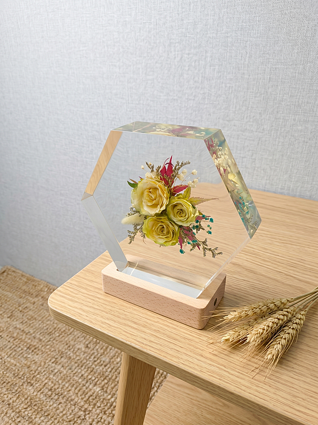 产品为六边形树脂干花摆件，包含六边形透明树脂块和原木底座，干花为三朵渐变黄绿色玫瑰搭配狗尾草、红叶和蓝绿色配草。保持产品主体物理结构绝对不变，不改变产品结构，产品中每张图只出现一次。树脂部分必须清亮透明犹如玻璃，画面干净无浮尘物。放置在浅原木纹理北欧风边桌上，背景是浅灰亚麻布艺墙面，角落放置天然亚麻地毯，北欧自然风空间。漫射柔和自然光，低饱和度色调，阴影清淡。三分法构图，微俯视拍摄。旁边搭配一小束天然干麦穗，增加自然生机。商业级4K摄影，画面通透干净，符合国内电商种草风格。保持3:4画面比例。负向词：西方面孔,金发,深眼窝,雀斑,杂乱房间,死黑阴影,硬光,乡村风,高对比度,欧美妆容,变形,扭曲,穿模,重叠,比例失调,浮尘,脏污,树脂浑浊,多个产品