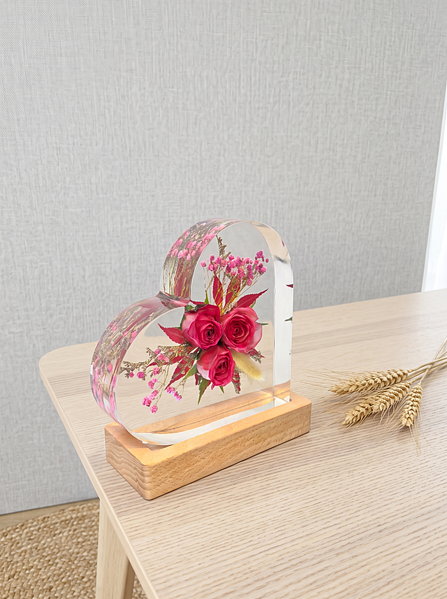 产品为心形树脂干花摆件，包含心形透明树脂块和原木底座，干花为三朵玫红色玫瑰搭配狗尾草、红叶和粉色满天星。保持产品主体物理结构绝对不变，不改变产品结构，产品中每张图只出现一次。树脂部分必须清亮透明犹如玻璃，画面干净无浮尘物。放置在浅原木纹理北欧风边桌上，背景是浅灰亚麻布艺墙面，角落放置天然亚麻地毯，北欧自然风空间。漫射柔和自然光，低饱和度色调，阴影清淡。三分法构图，微俯视拍摄。旁边搭配一小束天然干麦穗，增加自然生机。商业级4K摄影，画面通透干净，符合国内电商种草风格。保持3:4画面比例。负向词：西方面孔,金发,深眼窝,雀斑,杂乱房间,死黑阴影,硬光,乡村风,高对比度,欧美妆容,变形,扭曲,穿模,重叠,比例失调,浮尘,脏污,树脂浑浊,多个产品