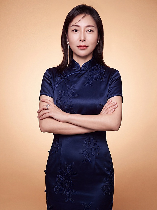 参考图中人物的脸型、发型、年龄、五官特征完全保留，换装为中式立领藏青色旗袍形象照。身穿藏青色中式立领旗袍，旗袍面料为高级真丝缎面质感，带有精致的暗纹刺绣花纹，立领设计典雅端庄，剪裁修身合体，展现东方女性优雅气质。保留原有精致耳环和戒指配饰。呈双手抱胸的站姿，姿态优雅自信，身体挺拔，肢体舒展自然。标准焦段50mm，f/4光圈，平视视角，镜头与人物视线齐平；人物占画面70%，人物居中构图，位于画面中心，为视觉聚焦点，半身照，左右留少量背景空间；头部与画面顶部保留5%空间；头部发型完整，头肩比例协调，人物结构准确。发梢微微修整，干练大气，主体锐利，背景光线均匀柔和，强化色彩对比的高级感。暖调米杏色纯色渐变背景，呈现均匀柔和质感，极简干净，温暖雅致。画质超高分辨率、细节清晰，现代精致感风格。肤质精细化处理，眼神直视镜头，目光坚定且澄澈，表情自然从容。肤质自然细腻，肤色健康均匀，哑光肤质清爽干净，保留真实皮肤细节。杰作，最佳画质，超高细节，写实摄影，中近景人像，皮肤质感自然清晰，柔光，正面补光，画面干净清新，高清皮肤细节；面部光比1:1.5，4800K中性光，微微修饰脸型，肤质哑光细腻，面部雾面柔光，轻透裸肌感，真实毛孔纹理。严格保留参考图人物的面部特征和发型。