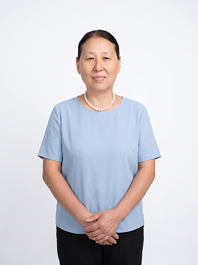 参考图中人物的脸型、发型、年龄、五官特征完全保留，生成专业商务形象照。标准焦段50mm，f/4光圈，平视视角，镜头与人物视线齐平；人物占画面70%，人物居中构图，位于画面中心为视觉聚焦点，半身照，左右留少量背景空间；头部与画面顶部保留5%空间，头肩比例协调，人物结构准确。身穿浅蓝色圆领短袖衬衫，面料呈细腻微微哑光质感；下装黑色长裤；着装整洁大方，气质端庄优雅，身形匀称，姿态端正，身体挺拔。呈自然站姿，双手在腹前自然交叠，肢体放松且自信。佩戴珍珠项链，保留配饰。发梢微微修整，干练大气，主体锐利。纯白色背景，均匀柔和质感，极简干净。画质超高分辨率、细节清晰，现代精致感风格。眼神直视镜头，目光温和坚定，表情自然，嘴角微微上扬。肤质自然细腻，肤色健康均匀，哑光肤质清爽干净，保留真实皮肤细节，符合中老年女性肤质特征。杰作，最佳画质，超高细节，写实摄影，中近景人像，皮肤质感自然清晰，柔光，正面补光，画面干净清新，高清皮肤细节；面部光比1:1.5，4800K中性光，肤质哑光细腻，面部雾面柔光，真实毛孔纹理。画面比例3:4
