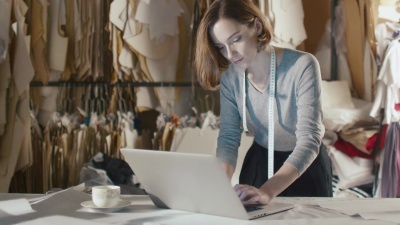 女服装设计师一边靠在工作室的桌子上一边在笔记本电脑上工作。