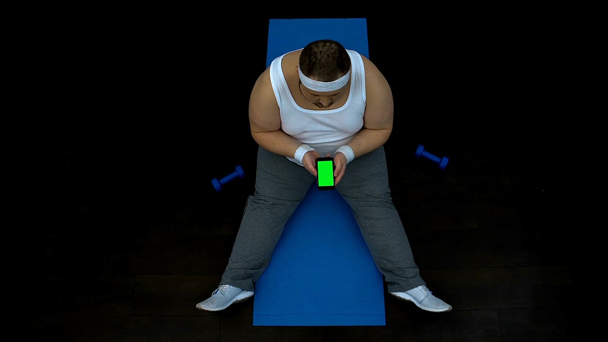 胖乎乎的男人在智能手机上使用健身应用程序，坐在垫子上，绿屏