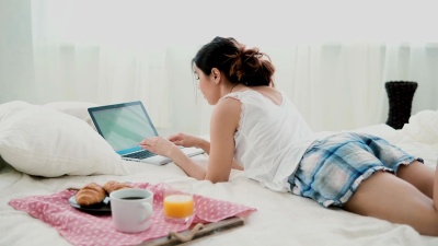 年轻女子在早餐时使用笔记本电脑，躺在家里的白色床上。深色头发的女孩在PC电脑上打字