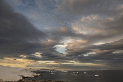 几座小冰山漂浮在倾斜的、白雪覆盖的土地旁边的水里，在被夕阳照亮的多云的天空下